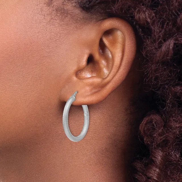 A person wearing a pair of textured oval hoop earrings in silver or white metal, shown from the side profile view.