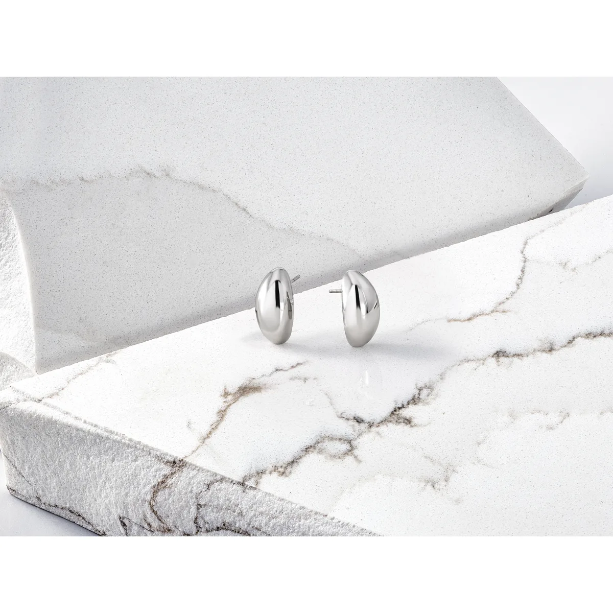A pair of polished silver oval stud earrings displayed on white marble and stone surfaces.