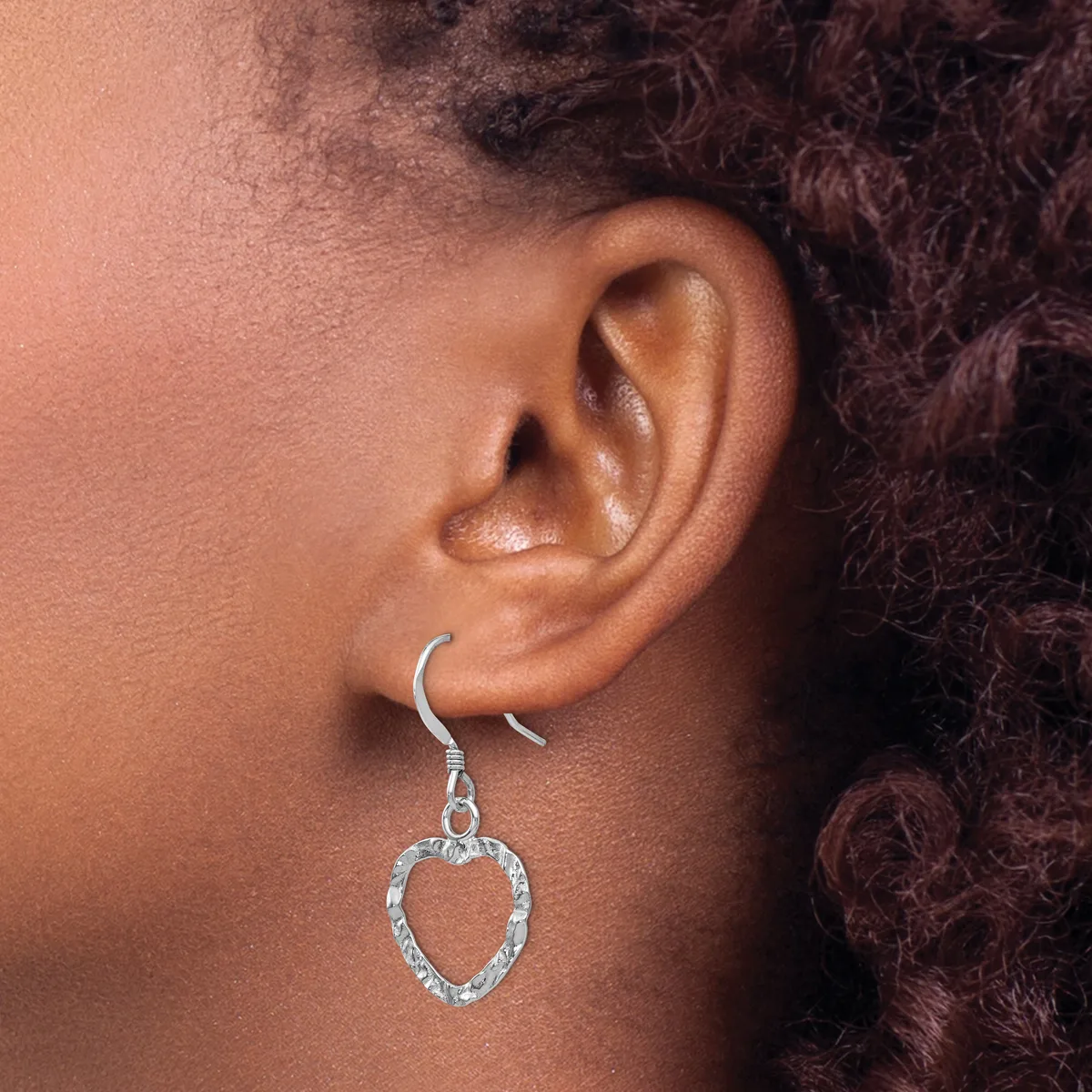 A person wearing a silver heart-shaped dangle earring with a textured finish on a fish hook wire, shown from the side profile of their ear.