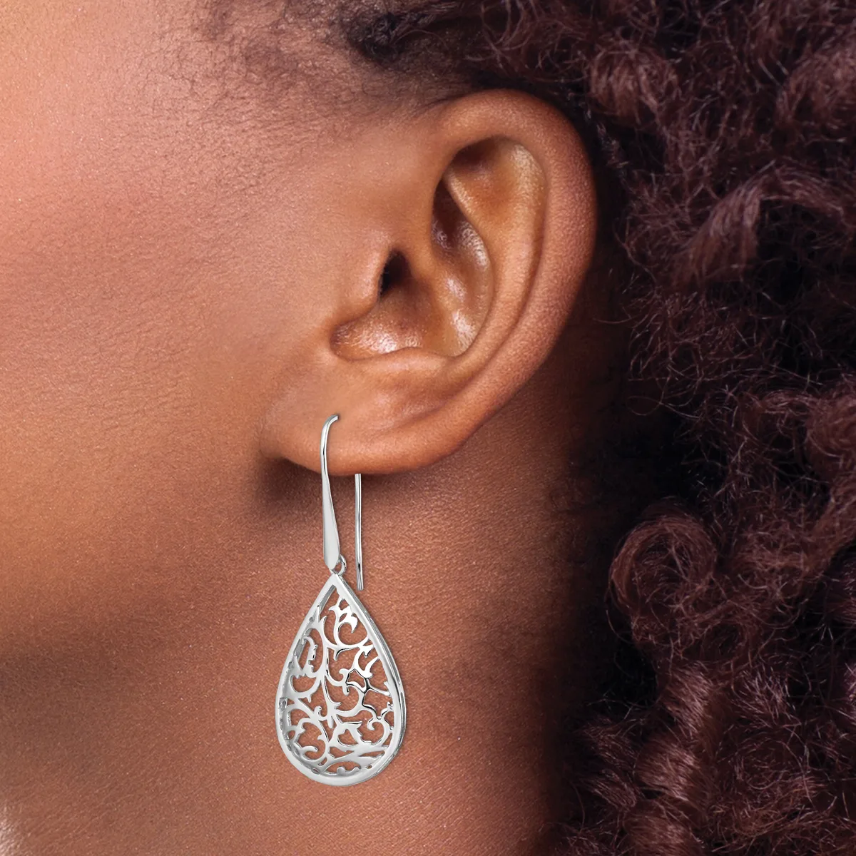A person wearing a silver teardrop-shaped filigree earring with an ornate scrollwork pattern, suspended from a curved hook.