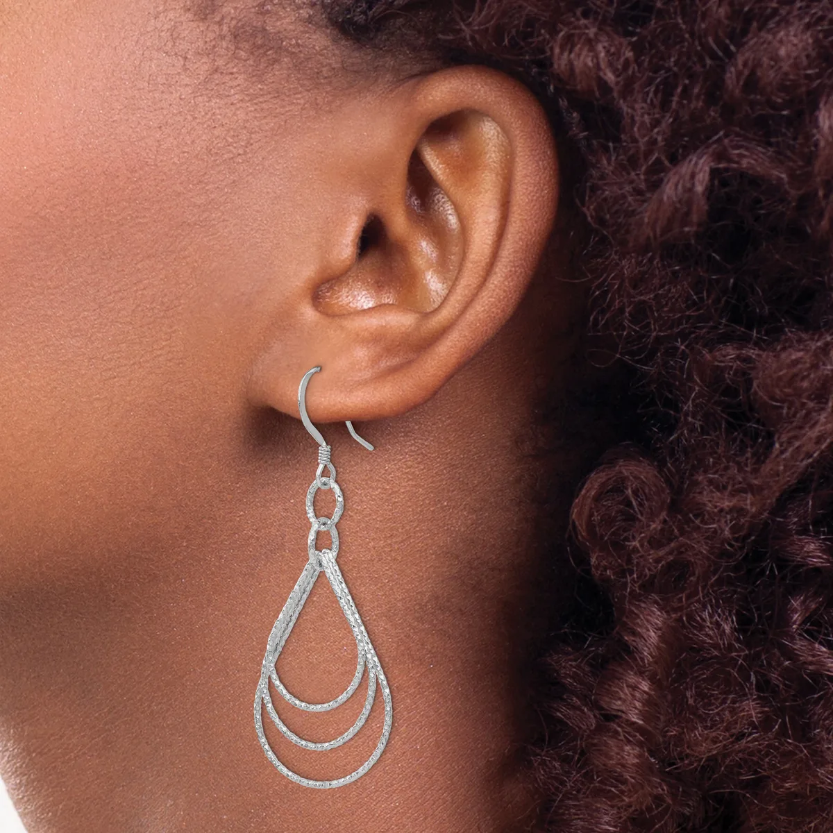 A person wearing a silver teardrop-shaped dangle earring with concentric nested loops, shown from the side against dark curly hair.