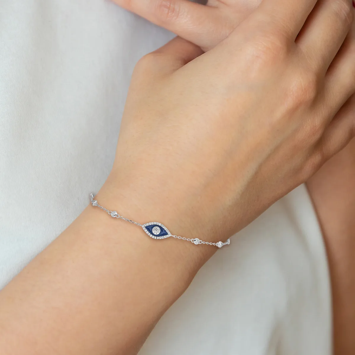 A delicate silver chain bracelet with a blue evil eye charm centered on the wearer's wrist, shown on a person wearing white clothing.