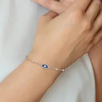 A delicate silver chain bracelet with a blue evil eye charm centered on the wearer's wrist, shown on a person wearing white clothing.