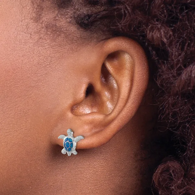 A person wearing a small turtle-shaped stud earring with blue stones and a sparkly finish in their earlobe.