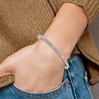 A person wearing a tan knit sweater and blue jeans displays a diamond tennis bracelet on their wrist.