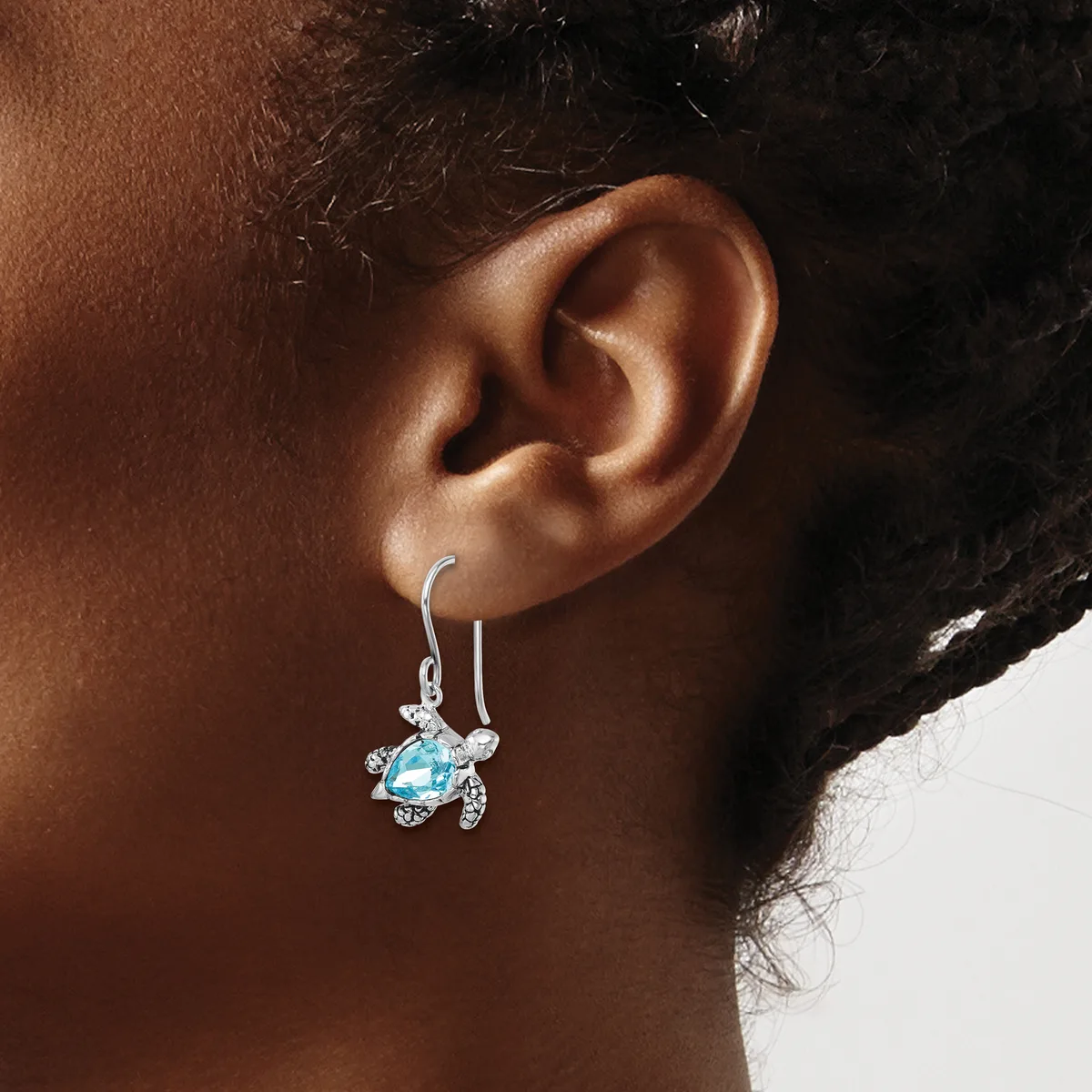 A person wearing a silver turtle-shaped earring with a blue gemstone body and rhinestone accents, displayed on a hook-style earring wire.