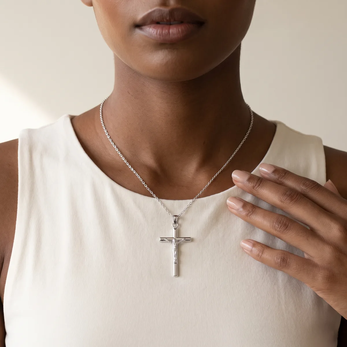 A person wearing a cream sleeveless top displays a silver cross pendant on a thin chain, with their hand raised near their chest.