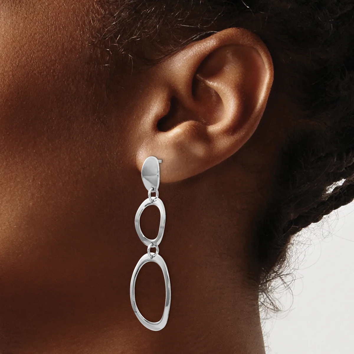 A person wearing a pair of silver-toned dangling earrings with two connected oval links hanging from a post-and-stud backing.