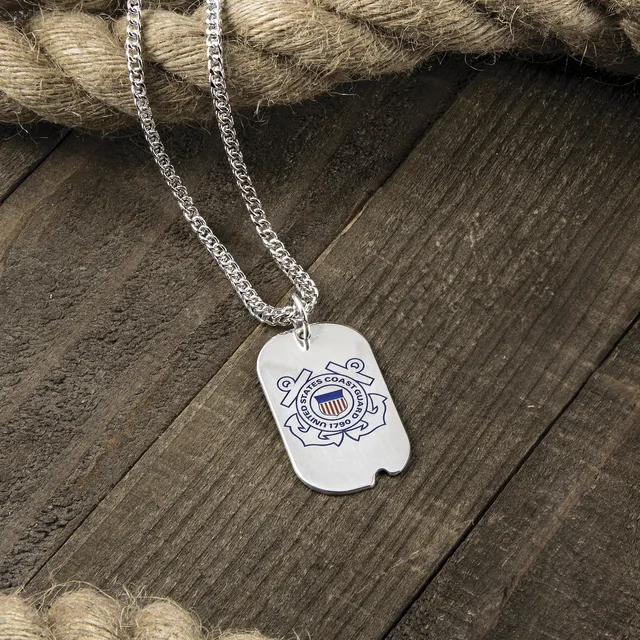 A silver dog tag pendant with the U.S. Coast Guard emblem engraved on it, hanging from a silver chain on weathered wood.