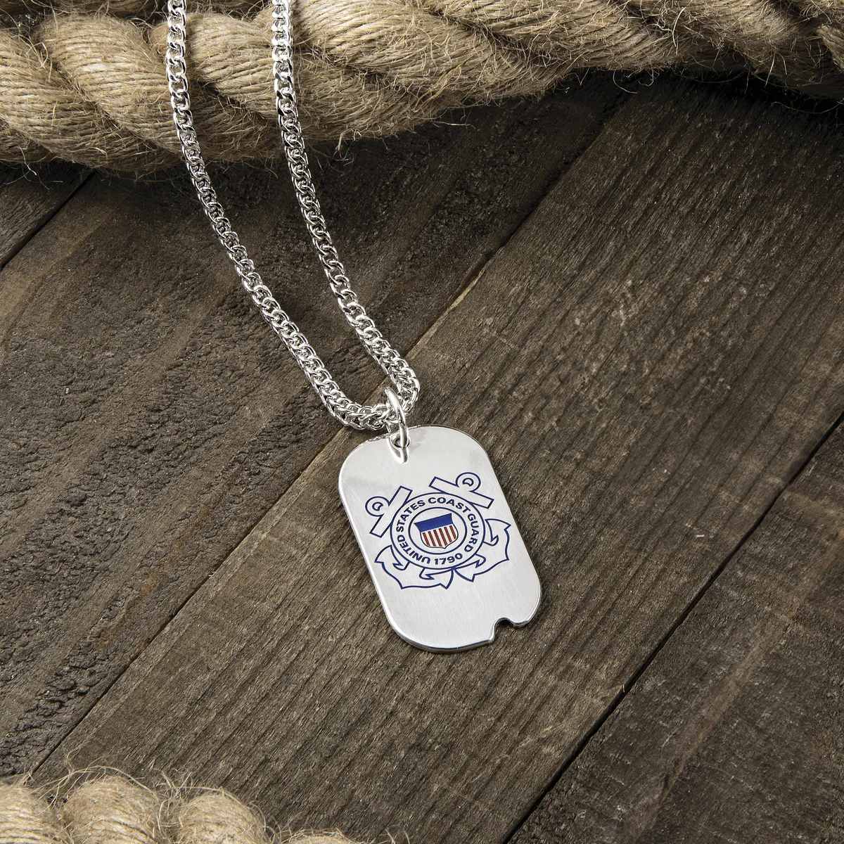 A silver dog tag pendant with the U.S. Coast Guard emblem engraved on it, hanging from a silver chain on weathered wood.