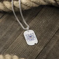 A silver dog tag pendant with the U.S. Coast Guard emblem engraved on it, hanging from a silver chain on weathered wood.