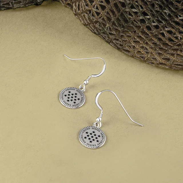 A pair of silver-toned dangle earrings with circular pendants featuring star designs and decorative beaded edges, displayed on a neutral beige surface next to dried plant material.