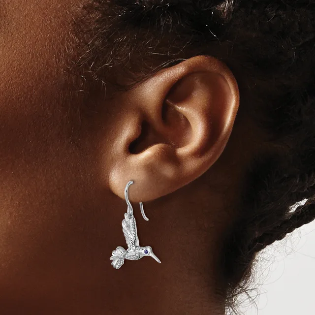 A person wearing a silver hummingbird-shaped dangle earring with gemstone details on a hook-style ear wire.