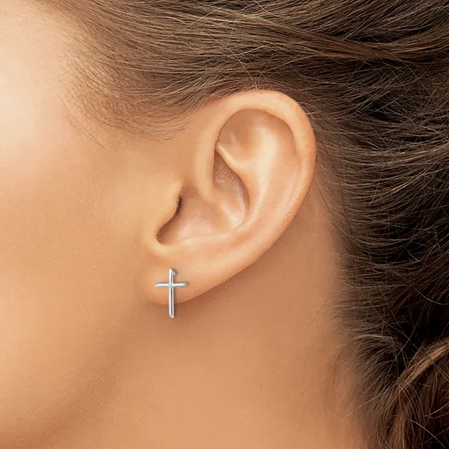 A silver cross earring stud is worn in the earlobe of a person with brown hair.