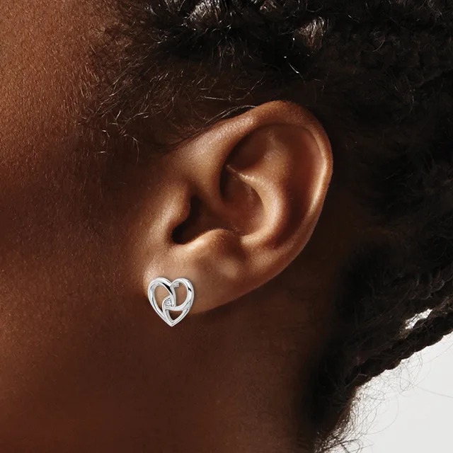A person wearing a small silver heart-shaped stud earring with an interlocking knot design in the center of the earlobe.
