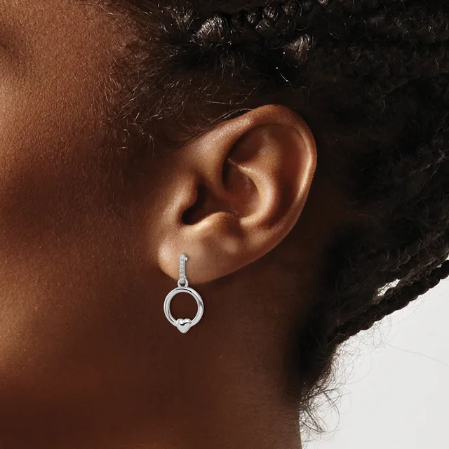 A silver drop earring with a circular pendant hangs from an earlobe, shown on a person with dark hair and brown skin.