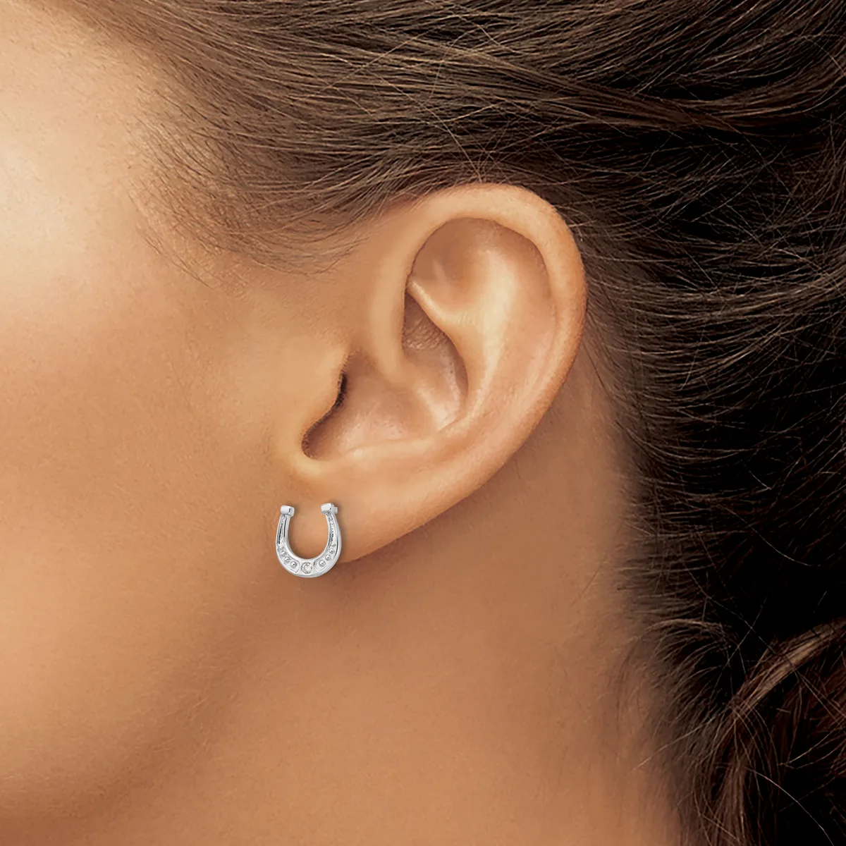 A person wearing a small horseshoe-shaped hoop earring set with small round stones in white metal, shown from the side view of their ear.