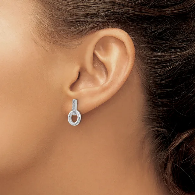 A woman wearing a small silver drop earring with a rectangular top section and oval loop bottom, set with small round stones.