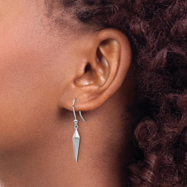 A person wearing a silver drop earring with an elongated pointed pendant hanging from a curved wire hook, shown from the side view against their ear.