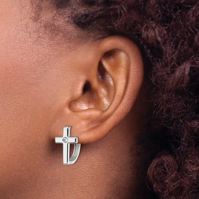 A silver cross earring with a polished finish and small diamond accent worn on the earlobe of a person with medium brown skin tone and dark curly hair.