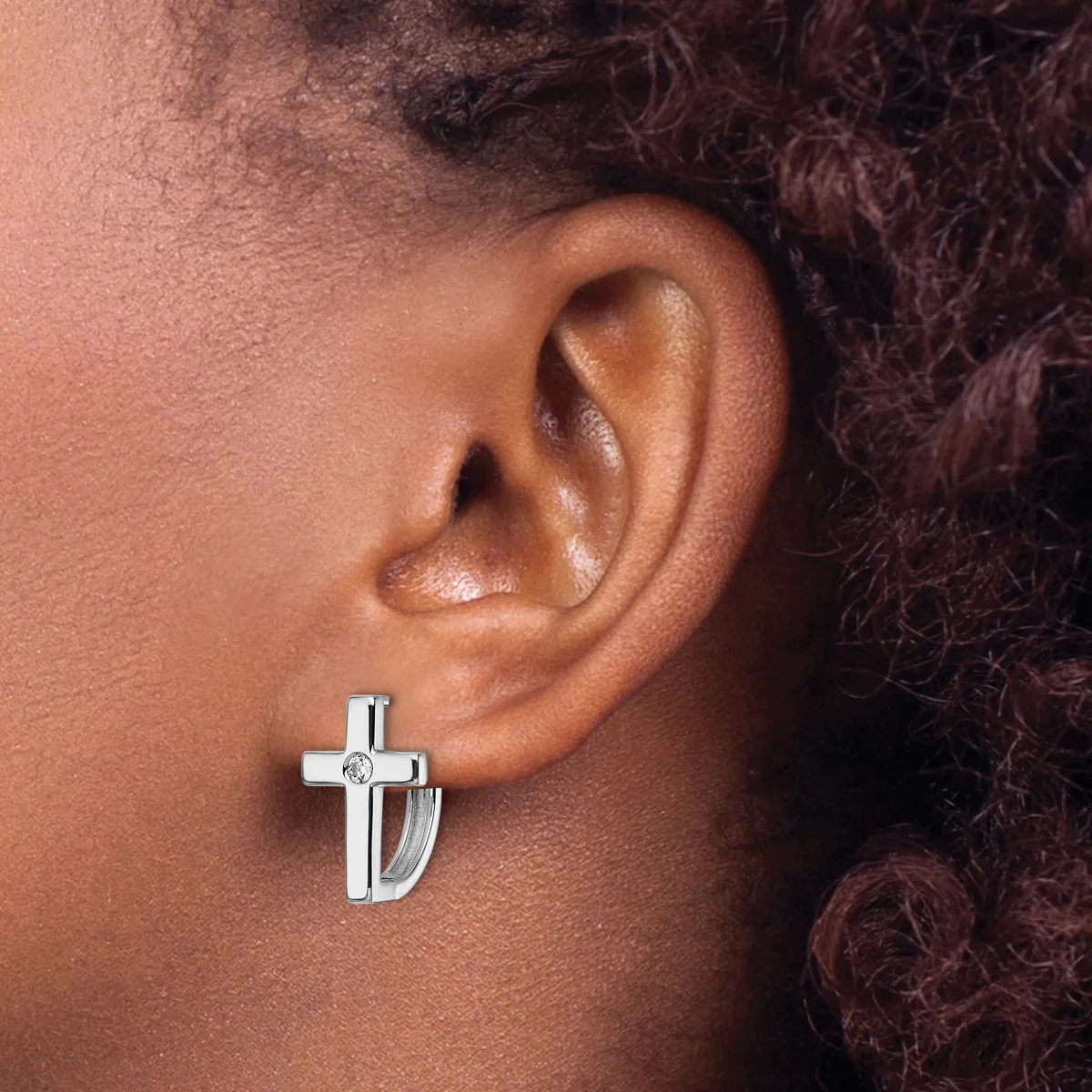 A silver cross earring with a polished finish and small diamond accent worn on the earlobe of a person with medium brown skin tone and dark curly hair.