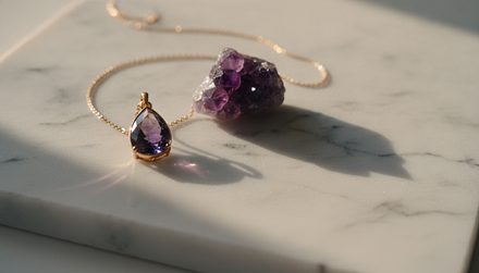 A gold chain necklace with a teardrop amethyst pendant lying on a white marble surface next to a rough amethyst crystal cluster.
