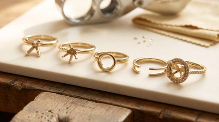 Five unset gold ring mountings on white marble — prong solitaire heads, a bezel rim, an open shank, and a diamond halo mounting — with loose melee stones and a jeweler's loupe behind.