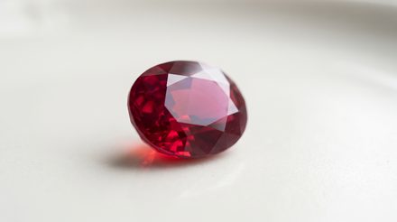 A single oval faceted ruby of deep vivid red resting on a pale porcelain surface, throwing a small pool of red light into its own shadow
