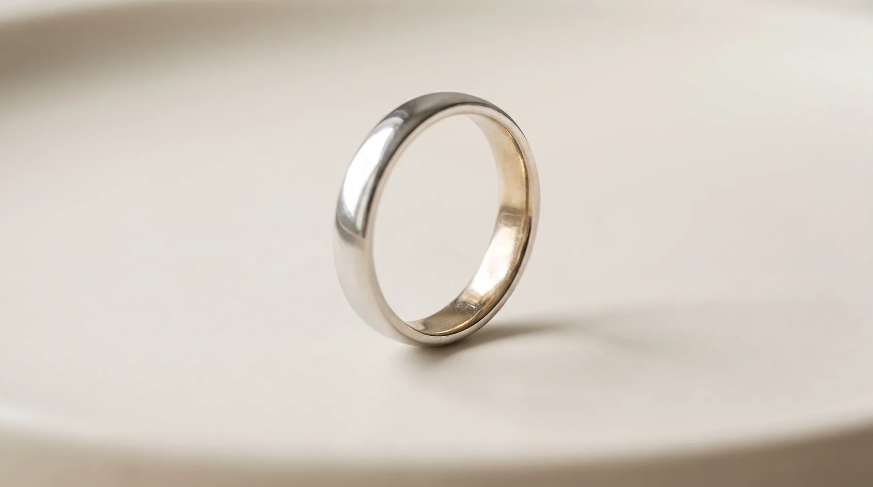 A plain white gold band standing on edge on a pale porcelain dish, its outer surface bright cool white while the inside curve of the shank shows a warm golden tone