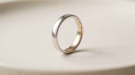 A plain white gold band standing on edge on a pale porcelain dish, its outer surface bright cool white while the inside curve of the shank shows a warm golden tone