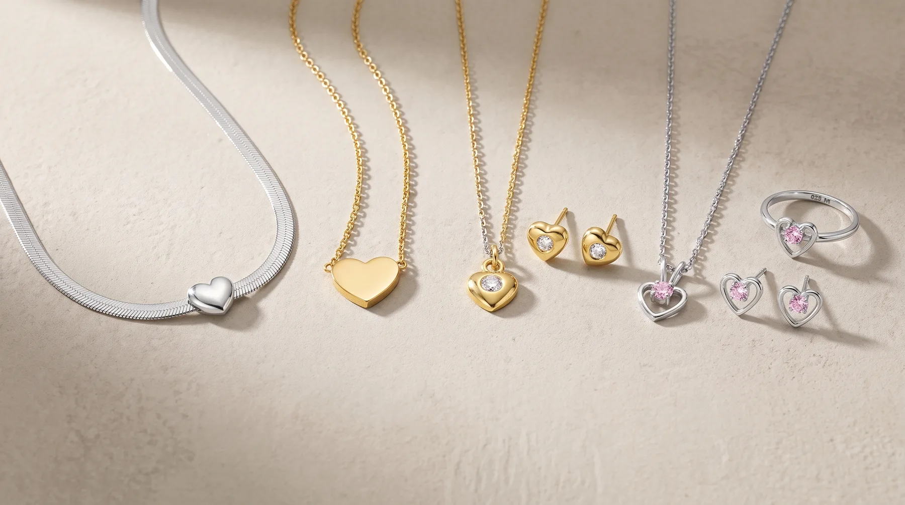 A collection of jewelry displayed on a neutral background, including silver and gold heart-shaped necklaces, matching earrings, and a ring with gemstone accents.
