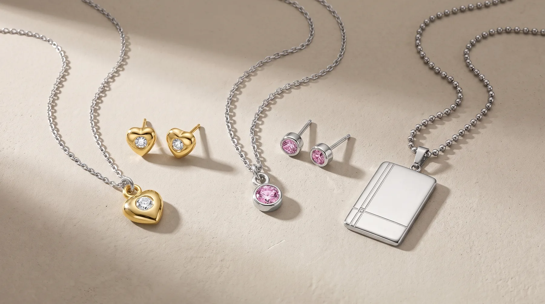 Three jewelry sets displayed on a beige surface: gold heart-shaped earrings and matching pendant necklace with clear stone, pink stone earrings and necklace, and silver rectangular pendant necklace with matching earrings.