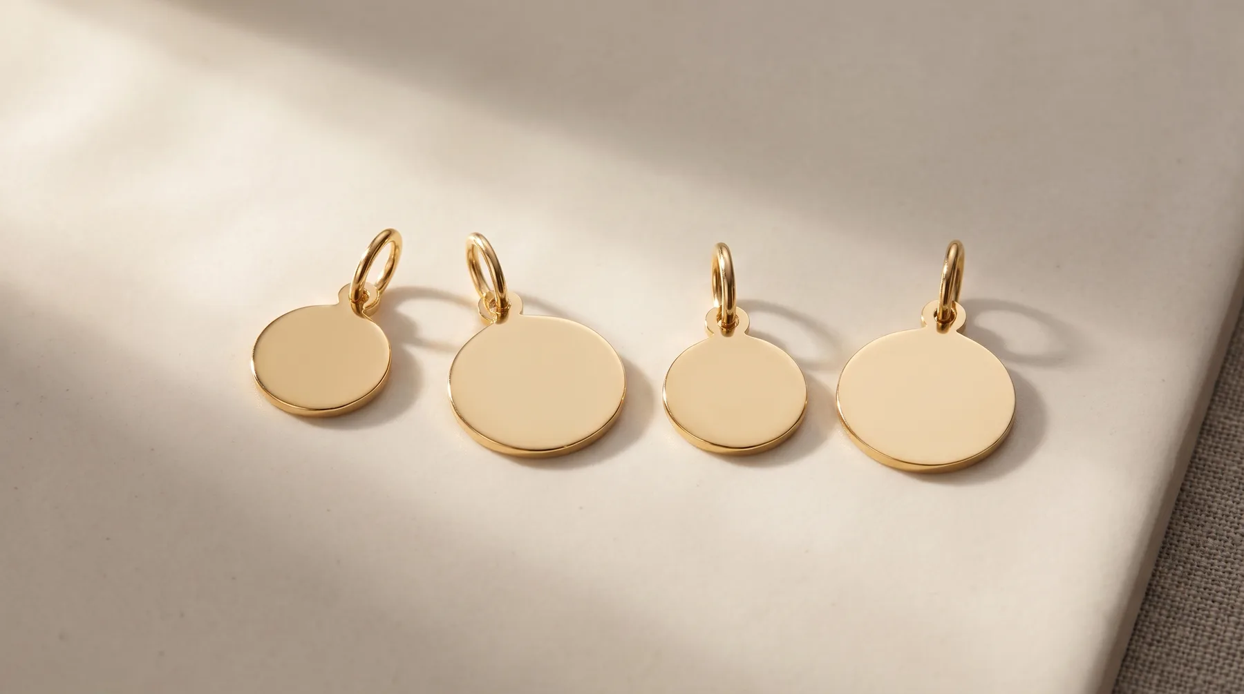 Four gold-colored round charm pendants with small loop attachments arranged in a row on a white surface, casting shadows.