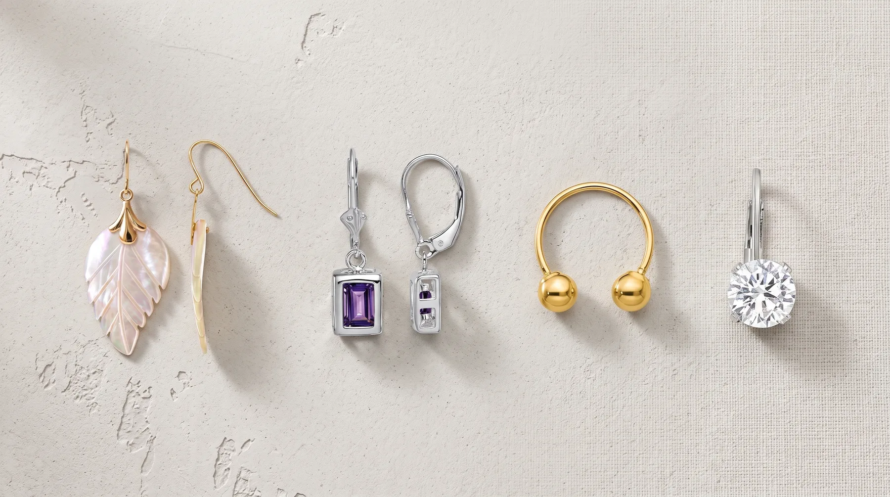 Five pairs of earrings displayed in a row on a textured beige surface: leaf-shaped drop earrings with gold wires, rectangular purple stone earrings with silver levers, gold ball hoop earrings, and round clear stone drop earrings with silver wires.