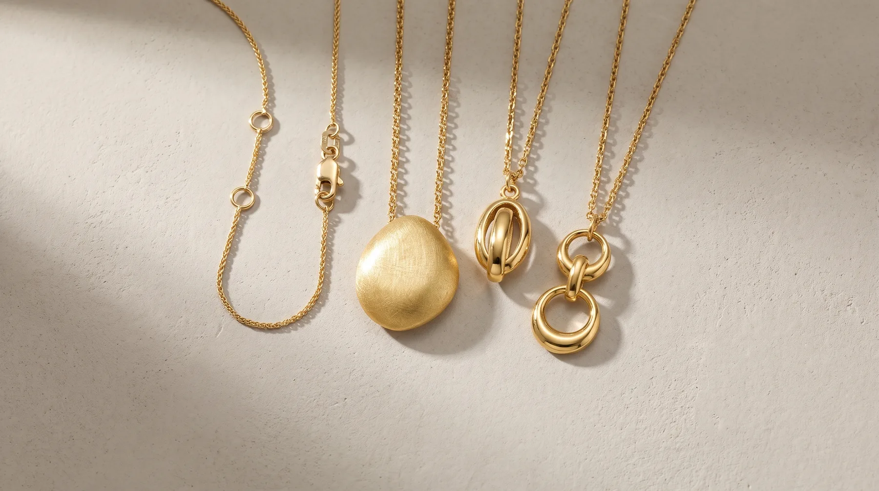 Five gold necklaces with various pendants arranged on a white surface, including a brushed dome pendant, interlocking rings, and chain details.
