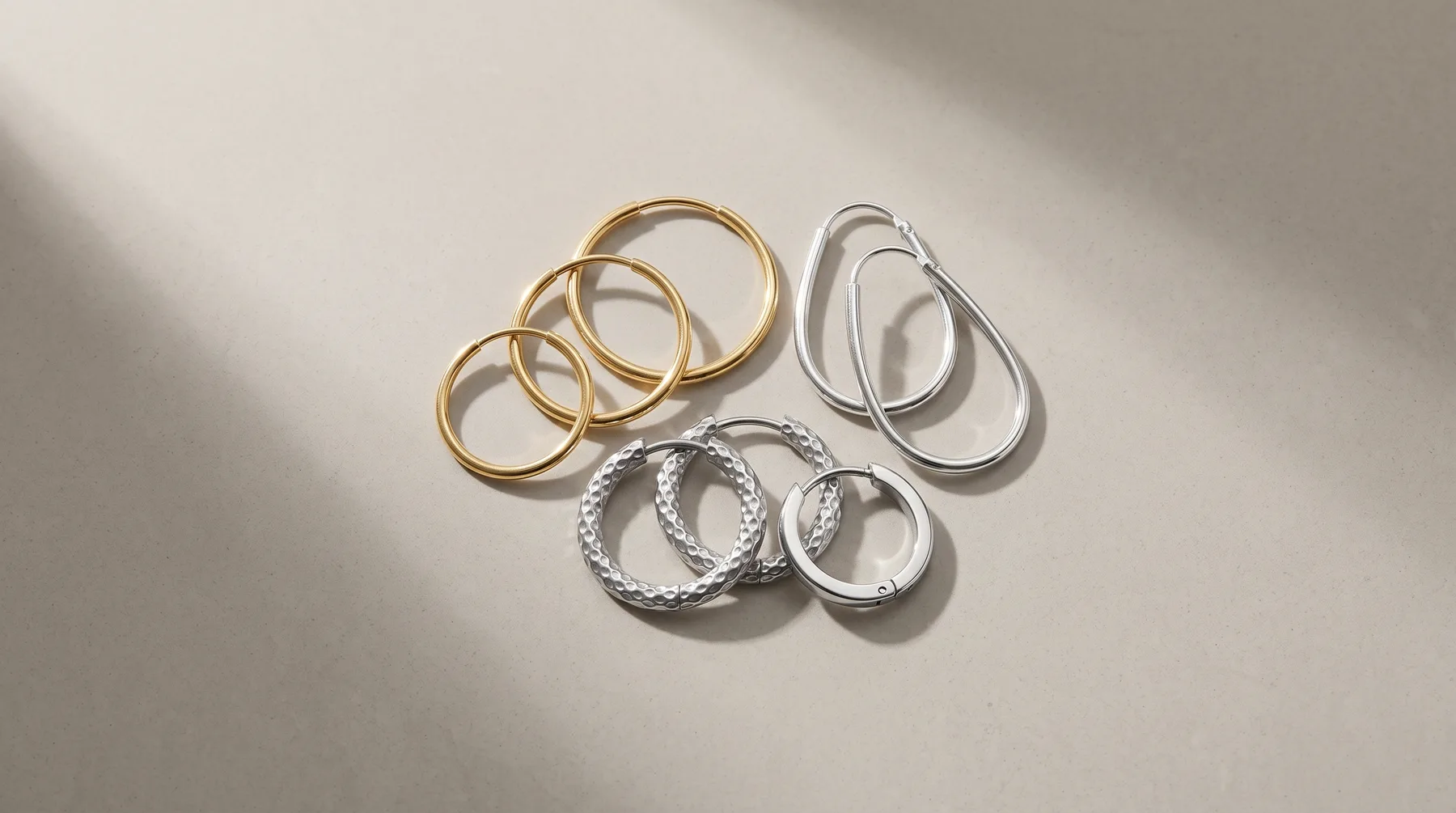 A collection of metal hoop earrings arranged on a white surface, including three gold-toned smooth hoops, two silver-toned smooth hoops, and two silver-toned textured hoops in various sizes.