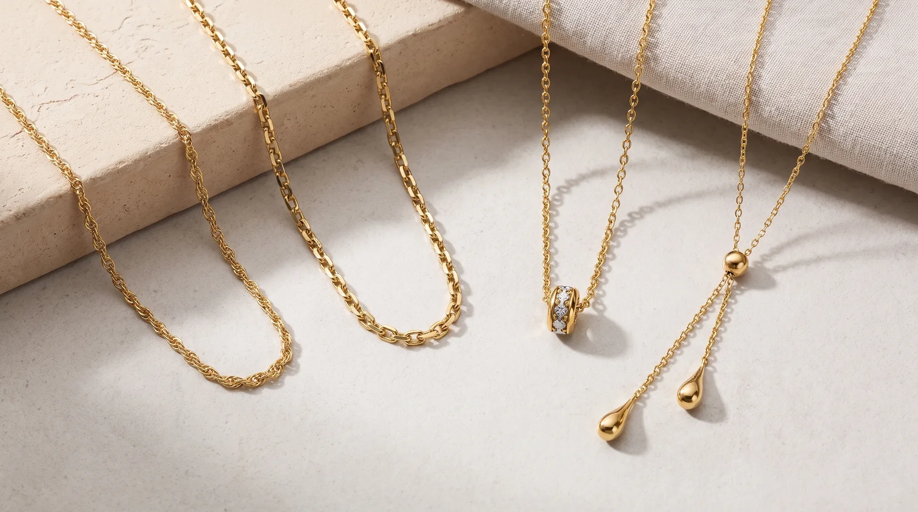 Five gold chain necklaces of varying styles displayed on a white surface with a beige block and cream fabric.
