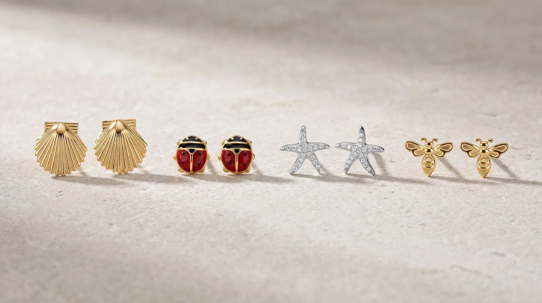 Four pairs of stud earrings arranged in a row on sand: gold shell-shaped studs, red and black ladybug-shaped studs, silver starfish-shaped studs, and gold bee-shaped studs.