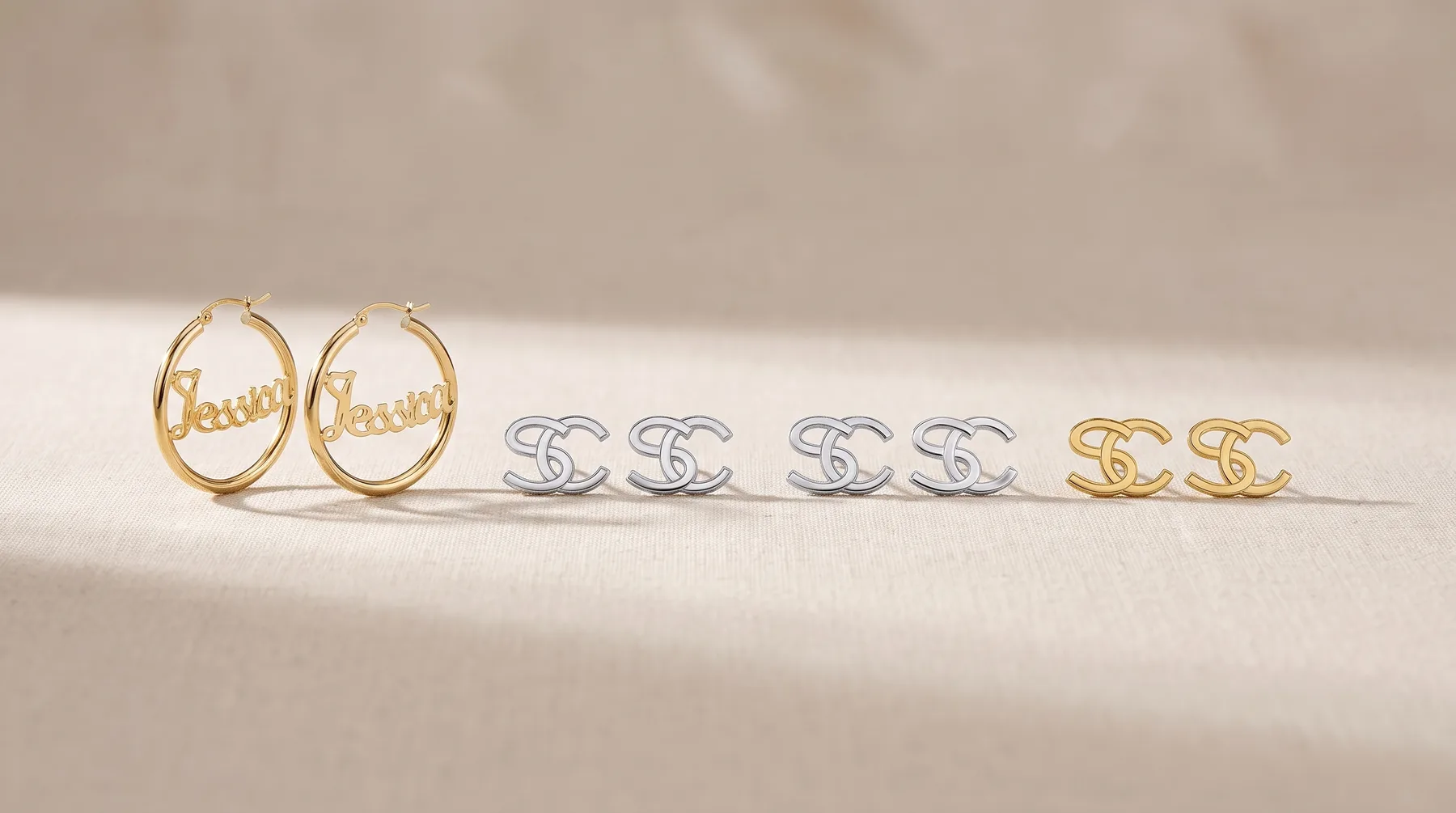 Six pairs of earrings arranged in a line on a beige surface: two gold oval hoop earrings with script text, four silver interlocking logo stud earrings, and two gold interlocking logo stud earrings.