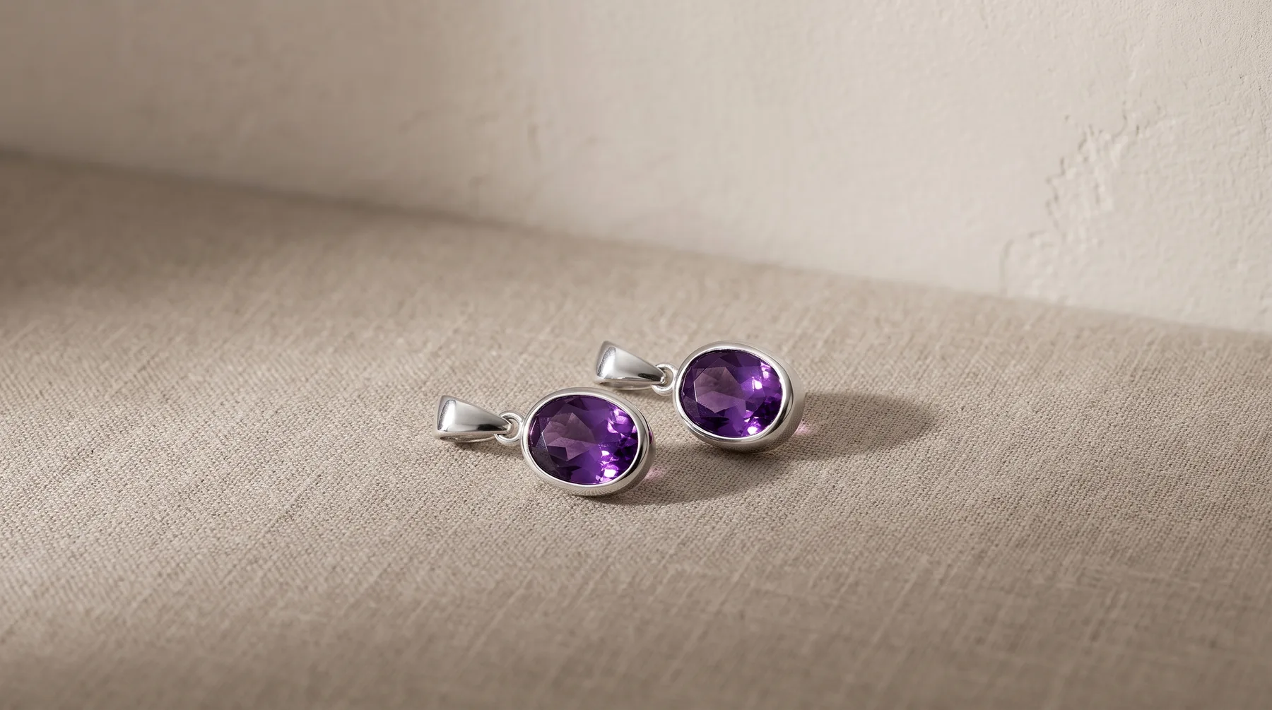 A pair of silver cufflinks with round purple amethyst stones set in polished bezels, displayed on a neutral beige fabric.