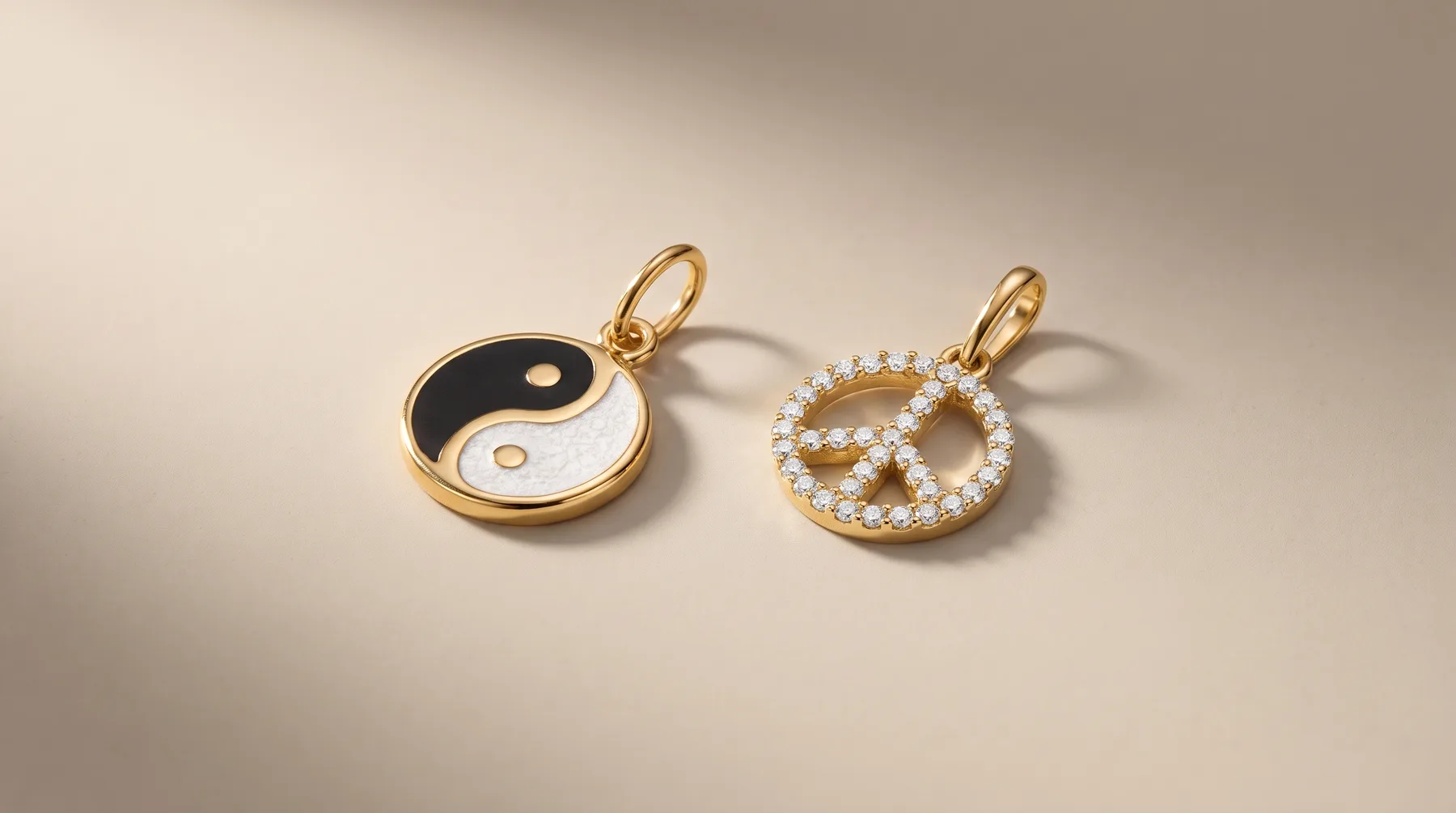 Two gold pendants with loops cast shadows on a light background: one featuring a black and white yin-yang design, the other a pretzel shape outlined in small diamonds.