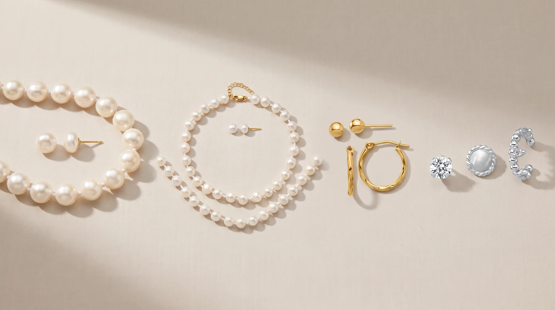 Pearl earrings, bracelet and necklace displayed with gold hoop earrings and diamond stud earrings on a neutral beige background.