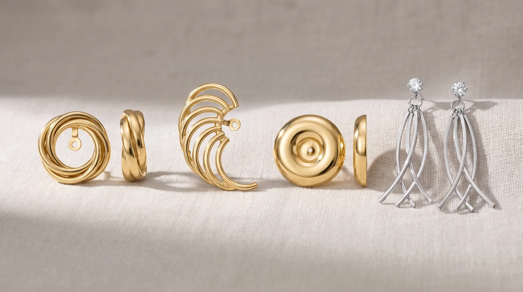 Six gold and silver earrings displayed on a white fabric surface, featuring circular hoops, twisted designs, disc shapes, and dangling geometric patterns with small gemstones.