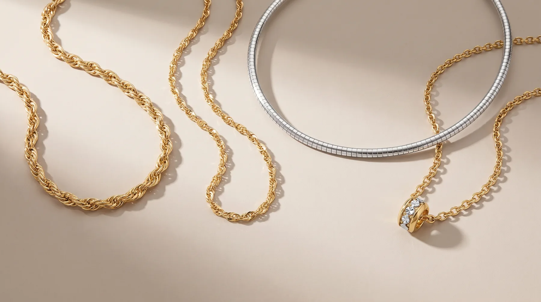 Five gold and silver chain necklaces of varying lengths and link styles arranged on a beige surface, including one with a small pendant charm.