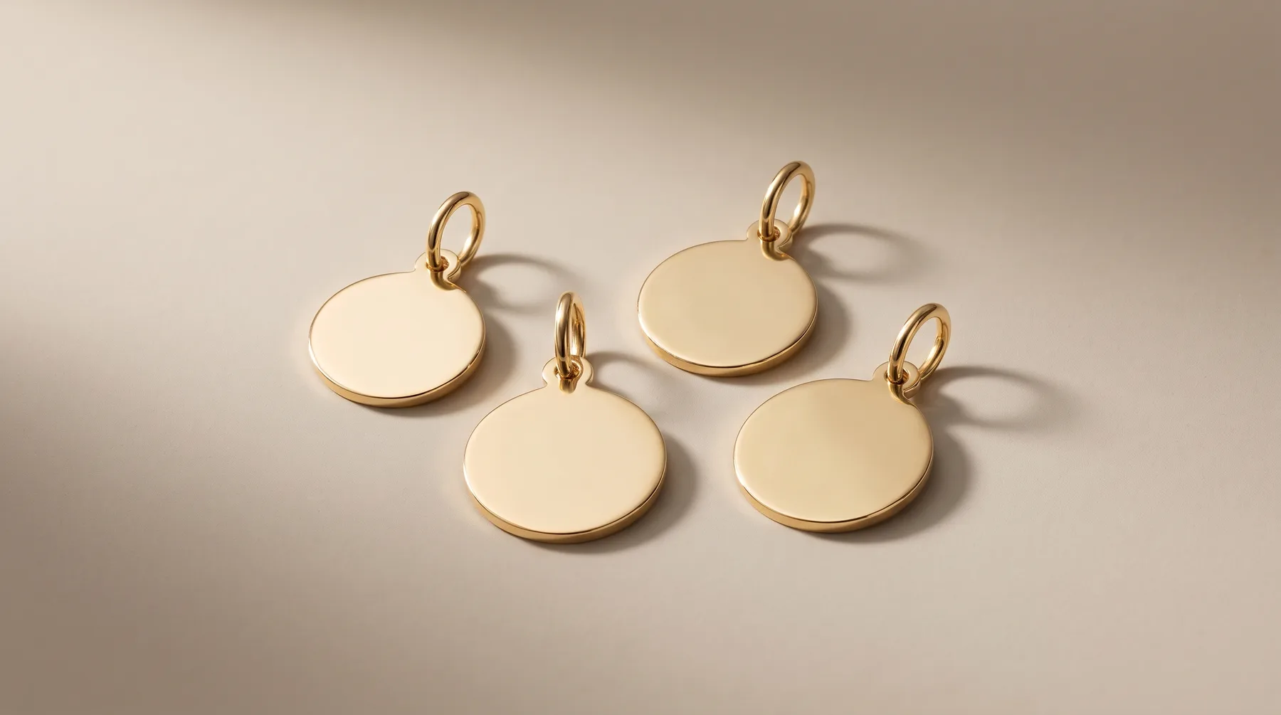 Four circular gold pendants with polished cream-colored enamel faces and gold loops, arranged on a neutral beige surface with shadows.
