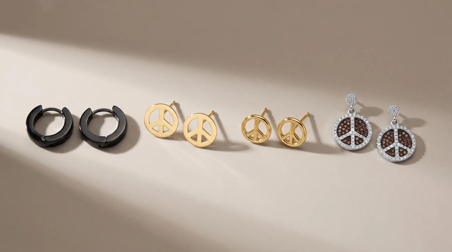 Five pairs of earrings arranged in a row, including black huggies, gold peace sign studs, two-tone peace sign studs, and silver peace sign drops with black and clear crystals.