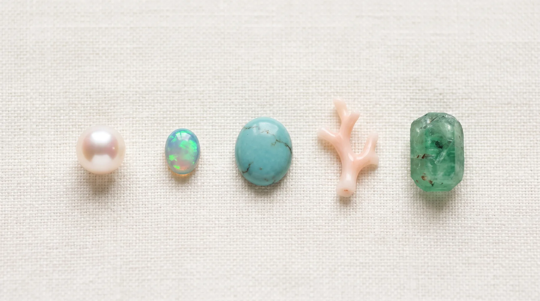 Five loose gems laid in a row on ivory linen: a white pearl, an opal flashing green and blue, a turquoise cabochon, a branch of pink coral, and a rough green emerald crystal