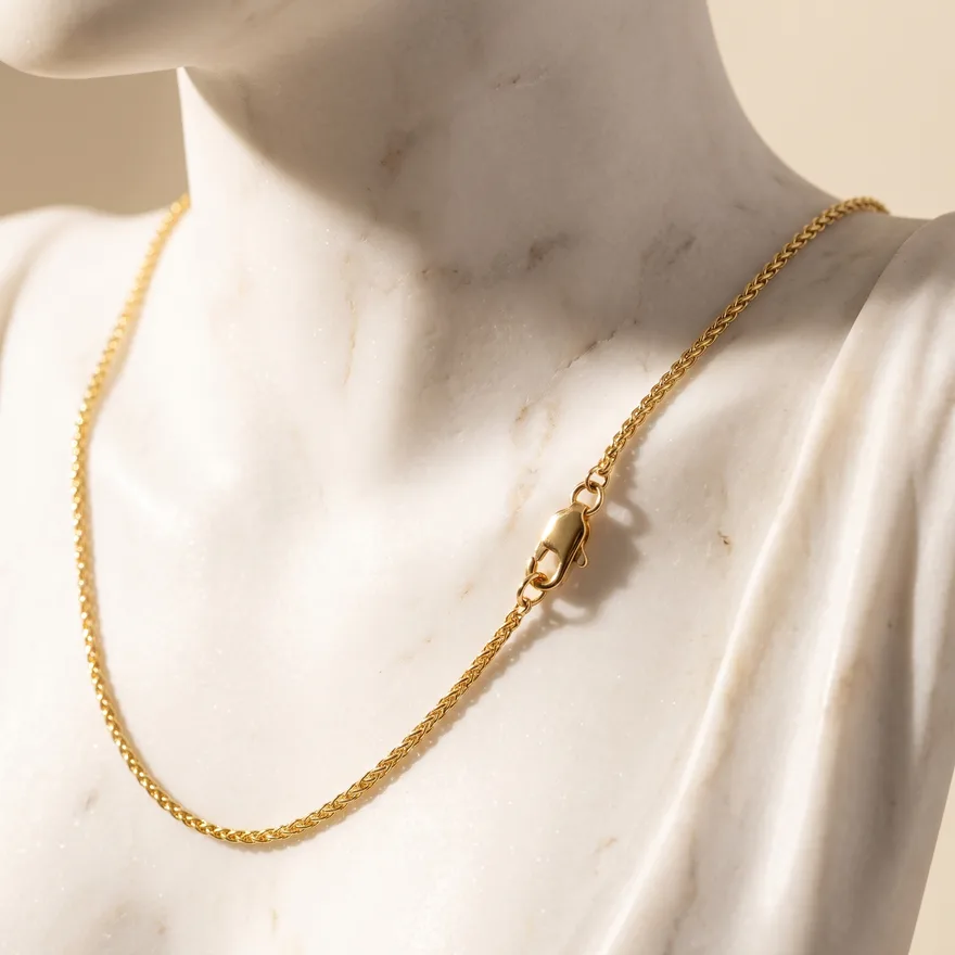 Gold wheat chain necklace draped over a white marble sculptural form in warm light