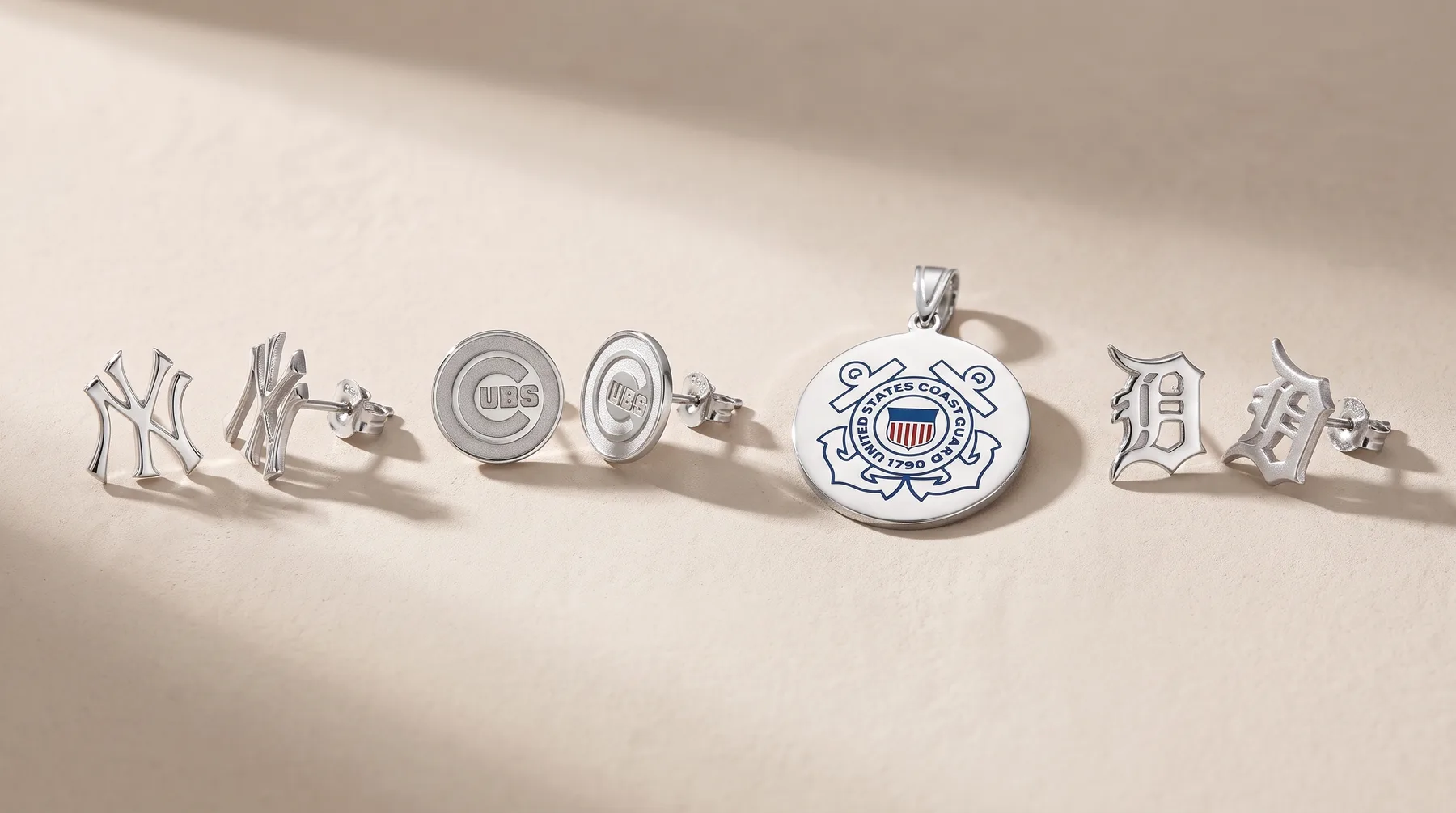 Six silver jewelry pieces arranged in a line, including cufflinks, stud earrings, a circular pendant, and a rectangular pendant with a stamped hallmark, all casting shadows on a light surface.