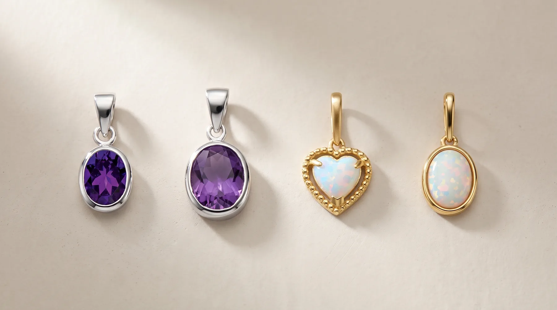 Four pendant necklace charms displayed in a row: two silver ovals with purple amethyst stones, one gold heart with opalescent stone, and one gold oval with opalescent stone.